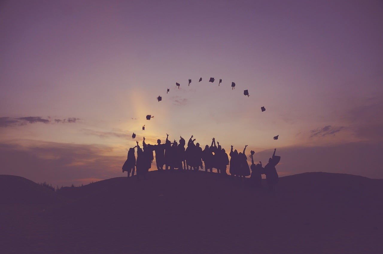Silhouetted graduates in stylish graduation outfits stand on a hill at sunset, joyfully tossing their caps into the air against a colorful sky.