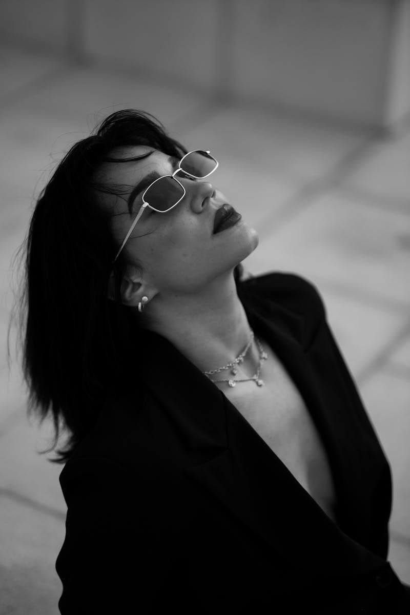 A black and white photo captures a person channeling celebrity-inspired looks with sunglasses and a blazer, eyes lifted. Their shoulder-length hair complements a chic necklace against the blurred, tiled background. A nod to fashion trends embraced by over-40 women.