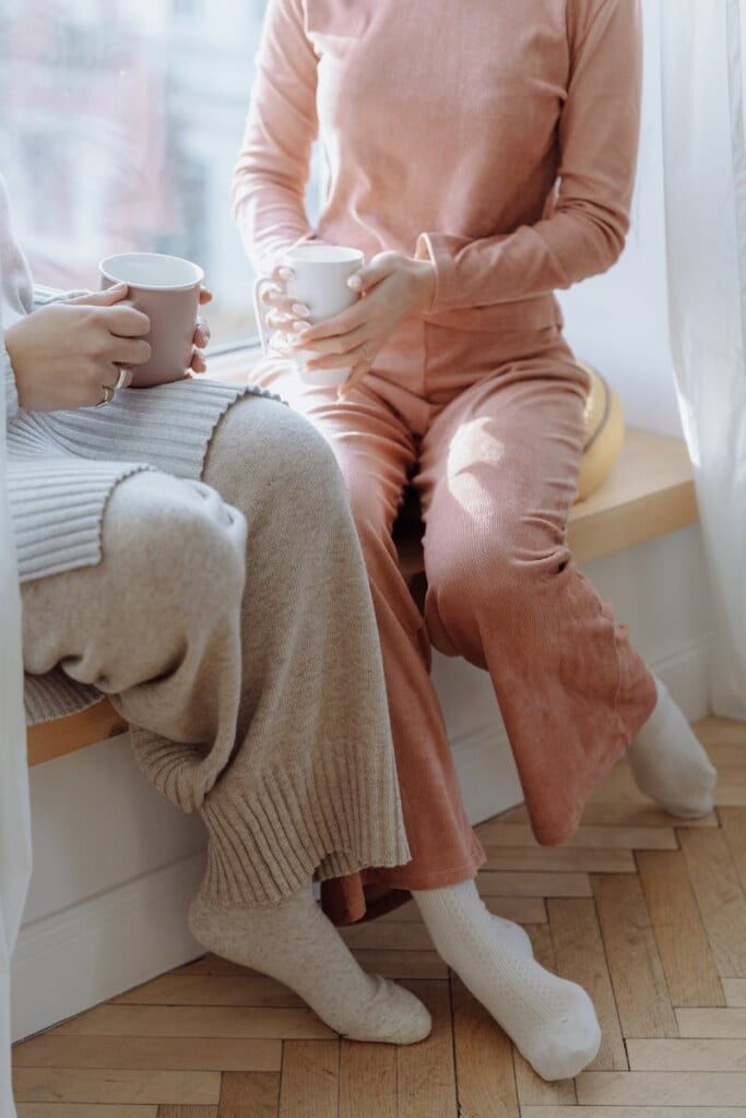 Two people sit on a window ledge in cozy outfits, holding mugs. One wears a chic beige sweater with matching pants, the other in a pink loungewear set. Natural light filters through sheer curtains, creating a warm, relaxed atmosphere perfect for working from home.