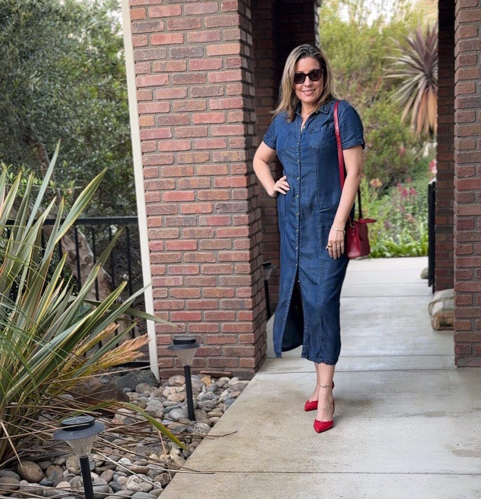 A woman in a denim dress and red heels stands on a concrete walkway next to a brick wall. She is wearing sunglasses and holding a red handbag. There are decorative plants and a garden in the background.