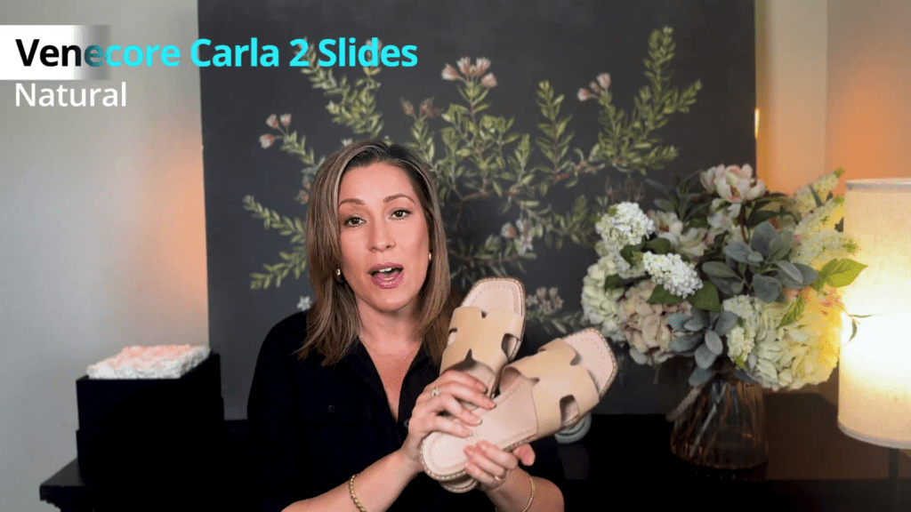 A person is holding a pair of beige slide sandals in front of a floral-patterned wall, showcasing their latest Amazon Haul. Text reads Venecore Carla 2 Slides Natural. A vase with flowers is in the background, lit by a lamp.